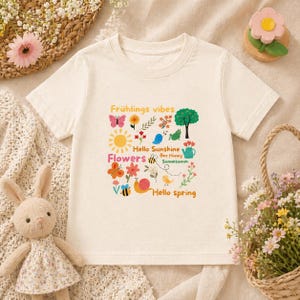 Könnte beinhalten: Cremefarbenes T-Shirt mit einem farbenfrohen Frühlingsmotiv. Das Design zeigt eine Sonne, Blumen, Schmetterlinge, eine Schnecke und die Sätze "Frühlings vibes", "Hello Sunshine", "Flowers" und "Hello spring".