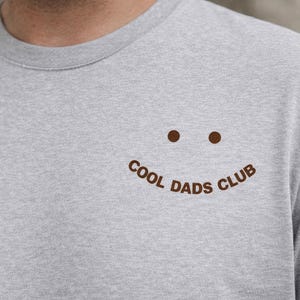 Könnte beinhalten: Hellgraues Rundhals-T-Shirt mit braunem Smiley-Gesicht und dem Text "Cool Dads Club". Das Shirt besteht aus einem weichen Material.