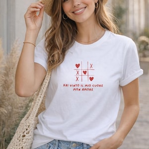 Könnte beinhalten: Weißes T-Shirt mit rotem Tic-Tac-Toe-Design mit Herzen und dem Text "HAI VINTO IL MIO CUORE MON AMORE." Das Shirt wird mit Jeans und einem Strohhut getragen.
