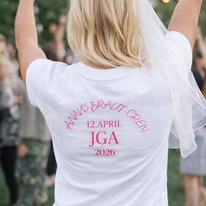 Könnte beinhalten: Ein weißes T-Shirt mit rosa Text, der lautet: "ANNA'S BRAUT GREN 12.APRIL JGA 2026". Eine Frau mit blondem Haar trägt das T-Shirt und hält ein Getränk. Ein weißer Schleier ist über ihrem Kopf drapiert.