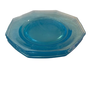 Può includere: Una pila di quattro piatti di vetro blu chiaro. Ogni piatto ha una forma ottagonale con un centro leggermente rialzato. I piatti sono di un colore blu pallido traslucido e sembrano essere vintage.