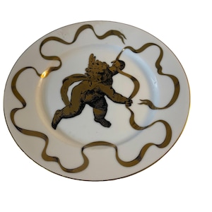May include: White plate with a gold rim, featuring a gold cherub figure and gold ribbon design. The cherub is depicted in motion, holding a ribbon. A decorative piece for display or dining.