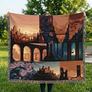 May include: A tapestry throw blanket featuring a landscape design with castles, arches, and a river. The color palette includes shades of brown, orange, and blue, with a fringed edge. The blanket is held up against a green background.
