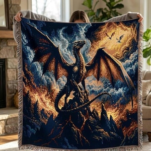 May include: A woven throw blanket depicting a dragon on a mountain with a fiery sky. The dragon is dark with orange wings, set against a blue and orange landscape. The blanket has a fringed edge. The design is detailed and colourful.