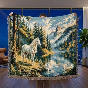 May include: A woven tapestry blanket depicting a tranquil landscape with a white horse by a lake. The scene includes trees, mountains, and a cloudy sky. The blanket has a fringed edge. The blanket is being held up by a person.