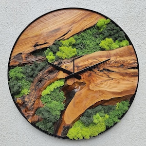 Peut inclure: Une horloge murale ronde avec un cadran en bois naturel et un cadre en métal noir. Le cadran de l'horloge présente une section de bois brun clair et foncé avec des accents de mousse verte. Les aiguilles noires indiquent l'heure.