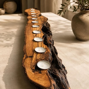 Puede incluir: Un portavelas largo y estrecho hecho de una sola pieza de madera natural con borde vivo. La madera presenta un patrón de veta prominente y corteza oscura y texturizada en el lateral. Contiene ocho pequeñas velas de té blancas y redondas.