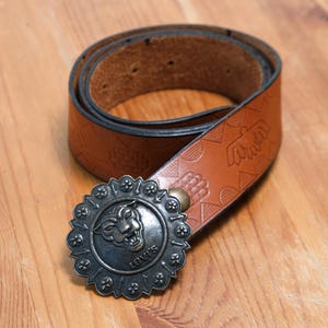 Può includere: Una cintura in pelle marrone con una fibbia decorativa color argento con un design a pantera. La cintura presenta motivi in rilievo e il logo Levi's sulla fibbia. La cintura è arrotolata su una superficie di legno.