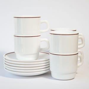 Könnte beinhalten: Weiße Keramikbecher und Untertassen. Die Becher haben ein schlichtes Design mit dunkelrotem Rand und einer passenden Linie am Boden. Die Untertassen sind gestapelt, und die Becher sind aufeinander gestapelt.