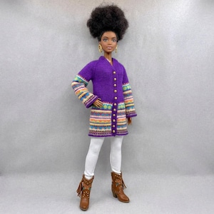 Könnte beinhalten: Puppe mit einem langen, lila Cardigan mit goldenen Knöpfen und bunten Ärmeln und Saum. Die Puppe trägt außerdem weiße Leggings und braune Cowboy-Stiefel mit Quasten. Die Puppe hat dunkle Haut, goldene Ohrringe und eine große Afro-Frisur.