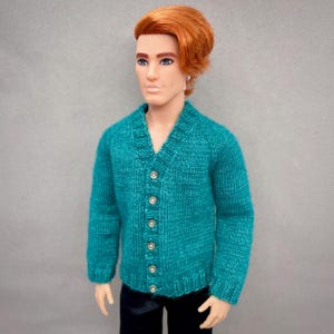 May include: A doll in a teal knitted cardigan with a V-neck and button closure, paired with black trousers. The doll has red hair and a small earring. The cardigan is a key piece of doll clothing.