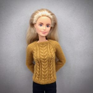 May include: A doll wearing a mustard yellow cable-knit sweater with long sleeves. The sweater has a textured pattern down the front and is paired with black pants. The doll has long blonde hair and is posed against a gray background.