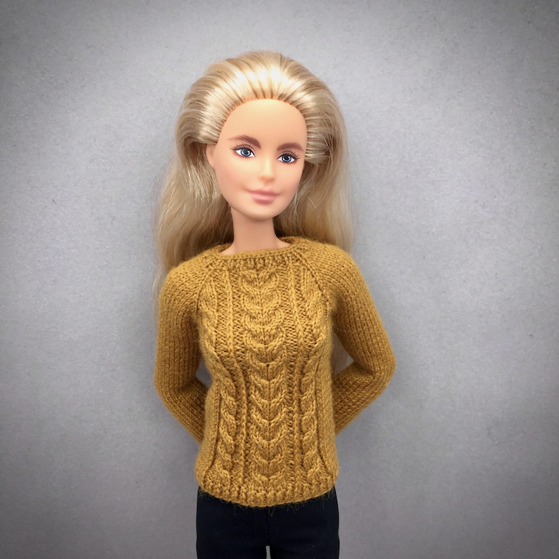 May include: A doll wearing a mustard yellow cable-knit sweater with long sleeves. The sweater has a textured pattern down the front and is paired with black pants. The doll has long blonde hair and is posed against a gray background.