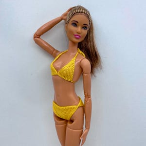 Puede incluir: Una muñeca con un bikini amarillo tejido. El traje de baño de dos piezas tiene un top halter y una braguita de cintura baja. La muñeca tiene el pelo largo castaño y posa con un brazo levantado.