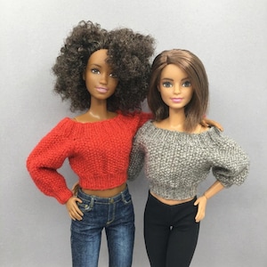 May include: Two dolls wearing off-the-shoulder sweaters. One doll is wearing a red sweater and blue jeans. The other doll is wearing a grey sweater and black trousers. Both dolls have light skin and brown hair.