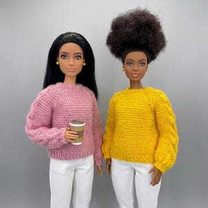 Op de afbeelding: Twee poppen dragen gebreide truien en witte broeken. Eén pop draagt een roze trui en houdt een koffiekopje vast. De andere pop draagt een gele trui. Beide poppen hebben donker haar en gouden oorbellen.