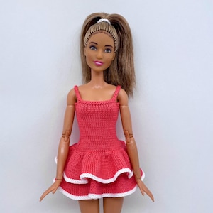 May include: A doll wearing a coral-colored knitted dress with white trim. The dress has a fitted bodice with thin straps and a flared skirt. The doll has long, light brown hair pulled back in a ponytail.