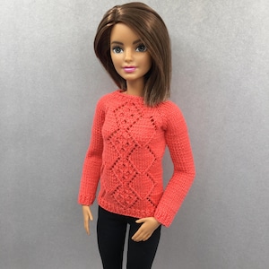May include: A coral-coloured sweater with a diamond pattern. The long-sleeved knit top features a textured design across the chest and upper sleeves. The doll is also wearing black trousers.
