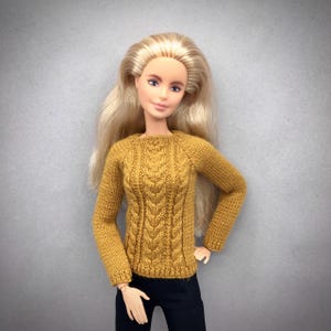 May include: A doll wearing a mustard yellow cable-knit sweater and black pants. The sweater has a crew neck and long sleeves. The doll has long blonde hair and is posed against a gray background.
