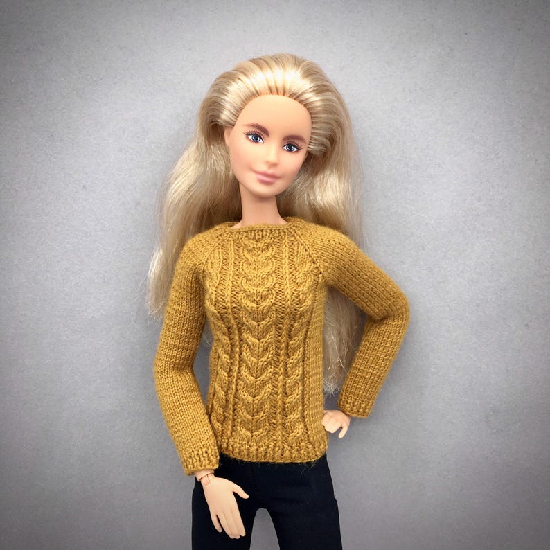 May include: A doll wearing a mustard yellow cable-knit sweater and black pants. The sweater has a crew neck and long sleeves. The doll has long blonde hair and is posed against a gray background.