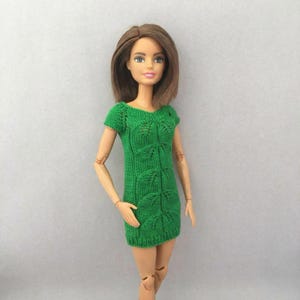 Può includere: Una bambola indossa un vestito verde a maniche corte lavorato a maglia con un motivo a foglie. L'abito ha uno scollo a V e una silhouette aderente. La bambola ha i capelli castani ed è su uno sfondo neutro.