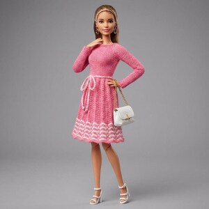 Puede incluir: Muñeca de moda con un vestido de punto rosa con un dobladillo estampado en blanco y rosa. La muñeca tiene cabello rubio, pendientes de aro dorados y un bolso blanco con una cadena dorada. También lleva sandalias blancas de tacón alto.