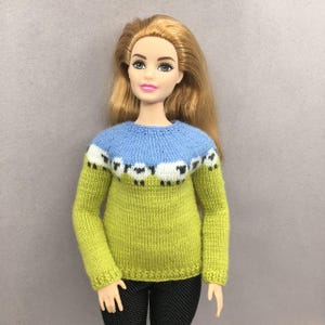 May include: A doll in a hand-knitted sweater. The sweater has a light blue yoke, a green body, and a band of white sheep with black faces. The doll has blonde hair and is wearing black trousers.