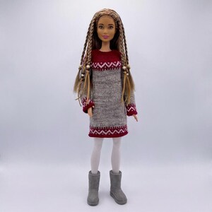 Pode incluir: Uma boneca vestindo um vestido de malha cinza e bordô com um padrão de zigue-zague branco. A boneca tem cabelo longo e trançado com contas de madeira, meias brancas e botas cinzentas. O vestido tem acabamentos bordô.