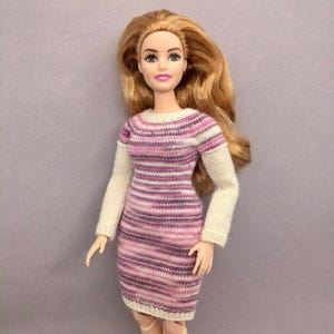 May include: A doll wearing a knitted dress with horizontal stripes in shades of pink and white. The dress has long white sleeves and a round neckline. The doll has long, wavy blonde hair and is posed against a solid gray background.