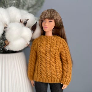 Könnte beinhalten: Eine Puppe in einem senfgelben Zopfmuster-Pullover. Der Pullover hat einen runden Ausschnitt und lange Ärmel. Die Puppe hat lange braune Haare. Im Hintergrund weiße Baumwollblüten vor grauem Hintergrund.