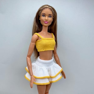 Puede incluir: Muñeca con top corto de punto amarillo y falda blanca escalonada con ribete amarillo. La muñeca tiene el pelo largo castaño y posa sobre un fondo blanco liso. El conjunto está hecho a mano.