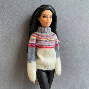 May include: A doll in a multi-coloured sweater with a grey turtleneck and sleeves. The sweater has a white lower section and black trousers. The doll has long black hair, set against a grey background.