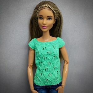 May include: A doll wearing a mint green, short-sleeved, knitted top with a leaf pattern. The doll has long brown hair and is wearing blue jeans. The top has a round neckline and is made of a soft, textured yarn.