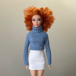 Op de afbeelding: Een pop met rood krullend haar, gekleed in een blauwe cropped coltrui en een witte minirok. De pop staat tegen een neutrale achtergrond. De trui heeft geribbelde details.