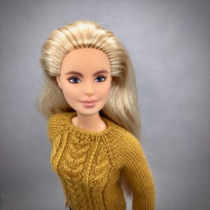 May include: A doll with long blonde hair and blue eyes wearing a mustard yellow cable-knit sweater. The sweater has a crew neck and a textured pattern. The doll's face is smooth with painted features.