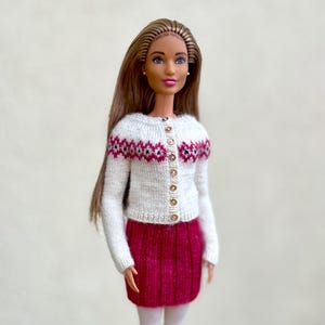 May include: A doll wearing a white knit cardigan with a geometric pattern in red and black, paired with a short, pleated, magenta skirt. The cardigan has gold-colored buttons. The doll has long, light brown hair and is wearing white tights.