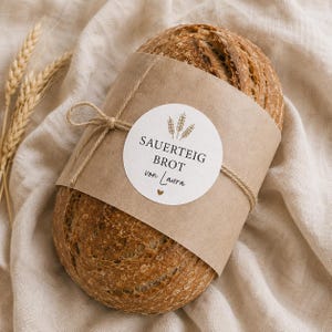 Könnte beinhalten: Ein runder Laib Sauerteigbrot, umwickelt mit einer braunen Papierbanderole und einer Kordel. Ein rundes weißes Etikett auf der Banderole zeigt den Text SAUERTEIG BROT von Laura, eine Grafik mit drei Weizenähren und ein kleines Herz.