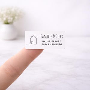 Könnte beinhalten: Ein weißes rechteckiges Etikett mit schwarzem Text und einer Strichzeichnung eines Hauses. Der Text lautet "FAMILIE MÜLLER", "HAUPTSTRABE 7" und "20144 HAMBURG". Das Etikett wird von einem Finger gehalten.