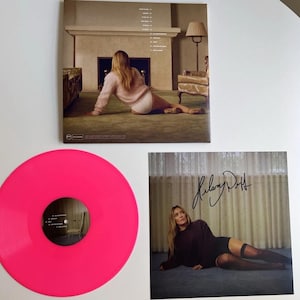 Pode incluir: Um disco de vinil rosa choque e duas capas de álbuns são exibidos. Uma capa apresenta uma mulher com um suéter rosa, a outra tem uma assinatura. O disco de vinil é de uma cor vibrante e sólida.