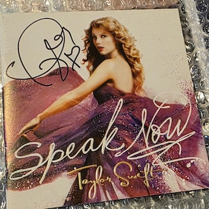 Puede incluir: Un CD "Speak Now" firmado por Taylor Swift. La portada del álbum muestra a Swift con un vestido morado, con el título del álbum en escritura blanca. La firma del artista está en tinta negra en la parte superior.