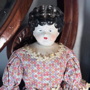 May include: A porcelain doll with a glossy black molded hairstyle, painted facial features including red lips and rosy cheeks, wearing a dress with a multi-colored circular pattern, puffed sleeves, and cream-colored lace trim at the neckline and cuffs.