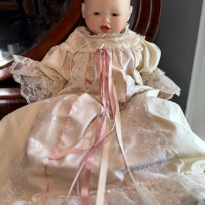 May include: A vintage porcelain baby doll with a serene expression. The doll wears a cream-colored dress with lace trim and pink ribbons. The doll's dress has lace sleeves and a lace-trimmed collar. The doll is a collectible item.