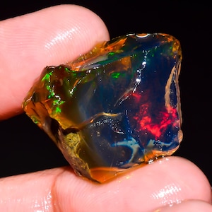 Op de afbeelding: Een levendige, ruw geslepen opaal edelsteen met een donkere basiskleur. De steen vertoont flitsen van rood, groen en blauw, met een doorschijnende kwaliteit. De opaal wordt tussen vingers gehouden, wat de natuurlijke, onbewerkte vorm laat zien.