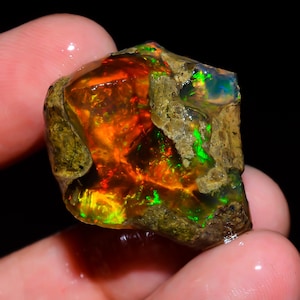 Könnte beinhalten: Ein roher Opal-Edelstein mit lebendigen Innenfarben. Der Stein zeigt feurige Orangen, Rot- und Grüntöne, mit einer dunklen, strukturierten Außenseite. Der Opal wird gehalten, was seinen natürlichen, unpolierten Zustand zeigt.