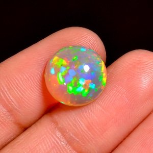 Puede incluir: Una piedra preciosa de ópalo redonda con una vibrante exhibición de colores, incluyendo verde, azul y naranja. La gema se sostiene en una mano, mostrando su superficie pulida y su juego interno de color. El ópalo tiene aproximadamente 2 cm de diámetro.