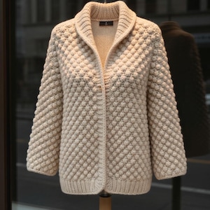May include: Cream-colored, textured knit cardigan with a shawl collar. The long-sleeved sweater features a unique bubble stitch pattern and is displayed on a wooden hanger. The cardigan is a neutral color, suitable for various outfits.