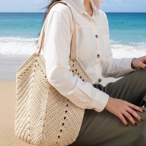 K&ouml;nnte beinhalten: Eine cremefarbene, geh&auml;kelte Tragetasche mit geometrischem Muster und Lederriemen. Die Tasche ist gro&szlig; und offen, geeignet f&uuml;r den Transport verschiedener Gegenst&auml;nde. Die Tasche wird an einem Strand gezeigt.