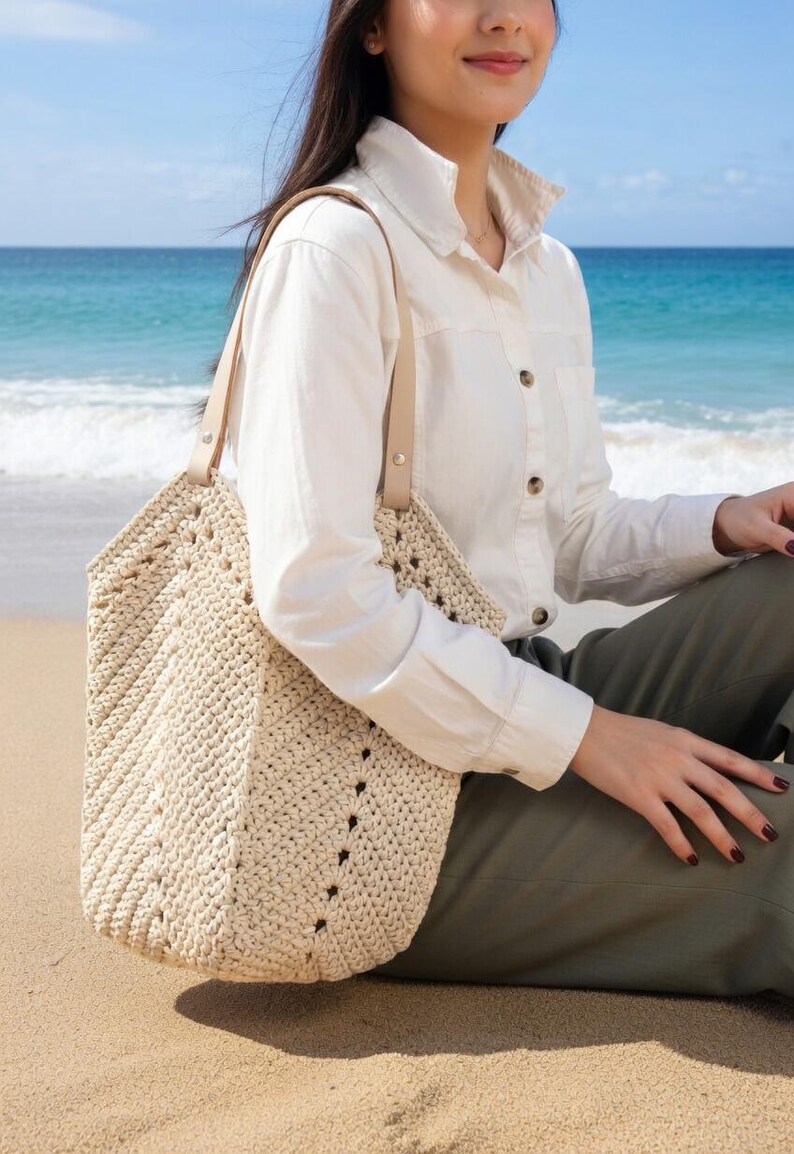 K&ouml;nnte beinhalten: Eine cremefarbene, geh&auml;kelte Tragetasche mit geometrischem Muster und Lederriemen. Die Tasche ist gro&szlig; und offen, geeignet f&uuml;r den Transport verschiedener Gegenst&auml;nde. Die Tasche wird an einem Strand gezeigt.