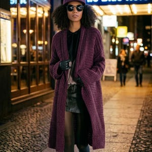 May include: A deep purple ribbed knit long cardigan worn over a black top and a black leather mini skirt, paired with black tights, black gloves, a matching purple baseball cap, and dark round sunglasses, standing on a cobblestone street.