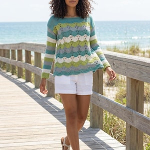 Puede incluir: Una mujer con un suéter de punto colorido con un diseño de vieira y mangas a rayas. El suéter presenta tonos de azul, verde, gris y blanco. Lleva pantalones cortos blancos y camina por un paseo marítimo de madera cerca de la playa.